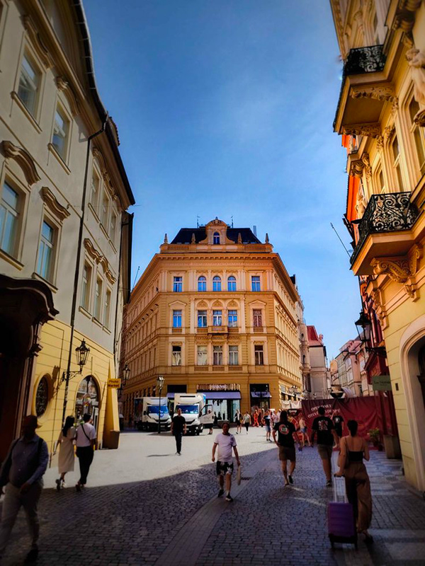 Widok ulicy z wąskiego brukowanego przejścia skierowanego w stronę dużego, ozdobnego budynku w stylu żółto-ochrowego pod czystym błękitnym niebem. Budynek ma klasyczną fasadę z dużymi łukowymi oknami na górnych piętrach i ciemnoniebieskimi detalami. Kilka białych samochodów dostawczych zaparkowanych na dole, w pobliżu sklepów. Ludzie spacerują po ulicy, a kobieta ciągnąc różową walizkę. Boki przejścia oprawiają niższe ściany sąsiednich starych budynków.
